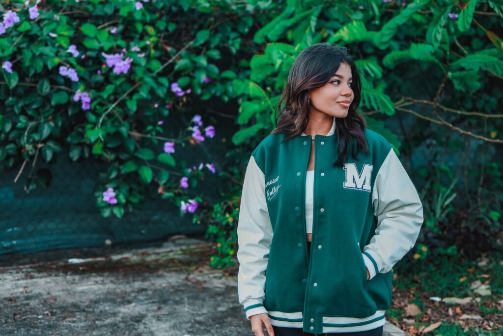 Charming young woman posing in a stylish green varsity jacket in an outdoor setting with lush foliage.